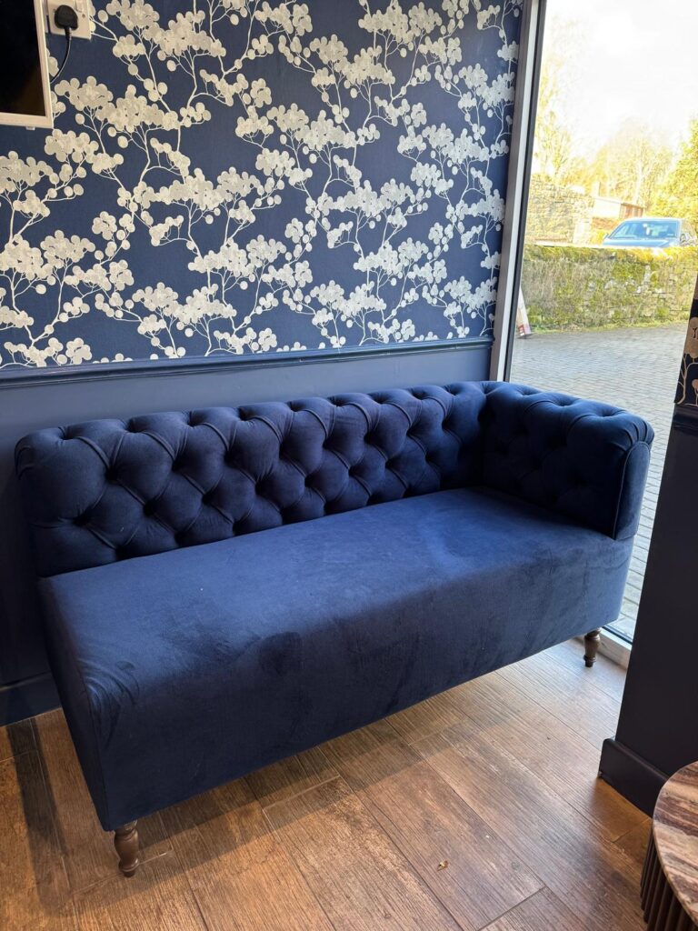 Deep blue sofa with a buttoned back and arms beside a window and floral wallpaper.