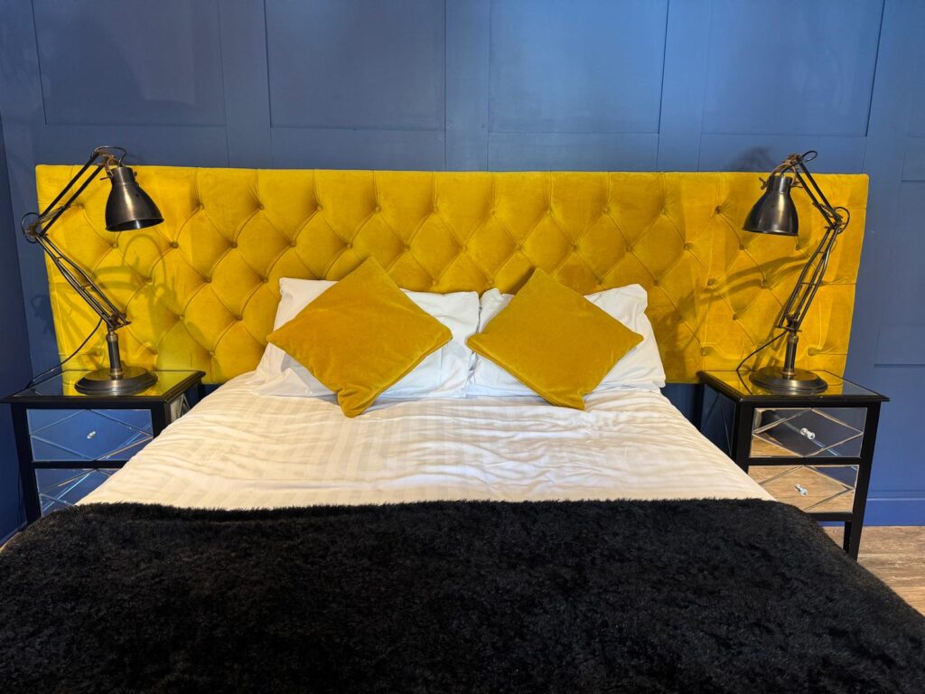 Front view of a wide mustard buttoned headboard, matching cushions and black lamps against a blue wall.