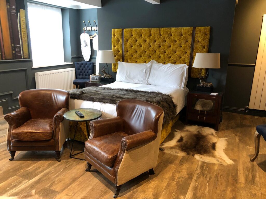 Bedroom with a tall mustard buttoned headboard, dark walls and a pair of brown armchairs.