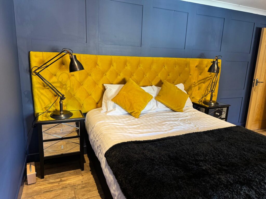 Wide mustard buttoned headboard and matching cushions against a blue panelled wall.