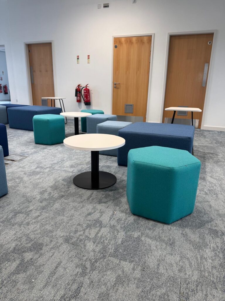 Teal hexagonal stool beside a white round table, with grey cubes and blue upholstered benches behind.