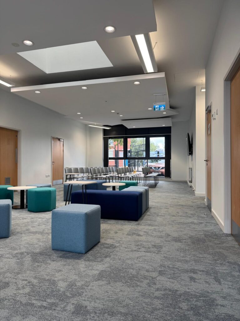 Long view through the room past grey cubes, navy benches and teal stools towards rows of chairs.
