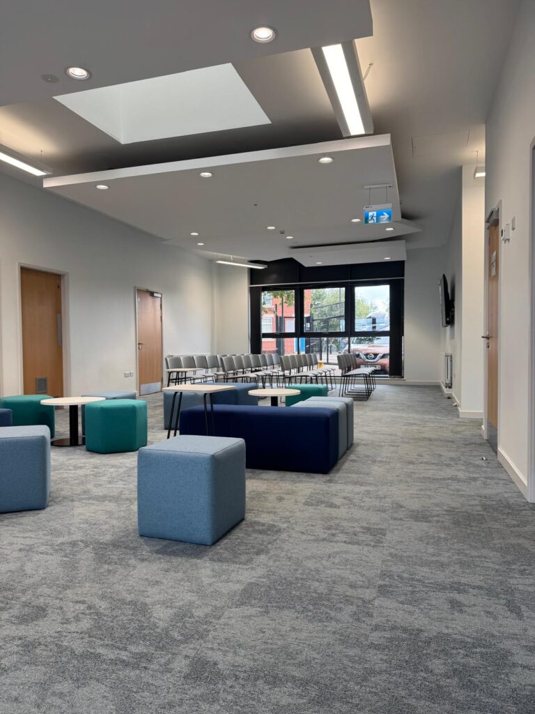 Grey cube in the foreground with a navy bench, teal stools and grey chairs beyond.