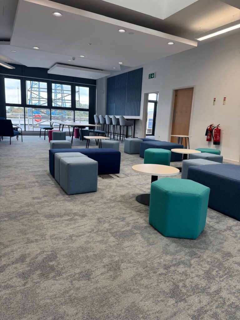 Wide view of blue, grey and teal soft seating, pale tables and high stools beside large windows.