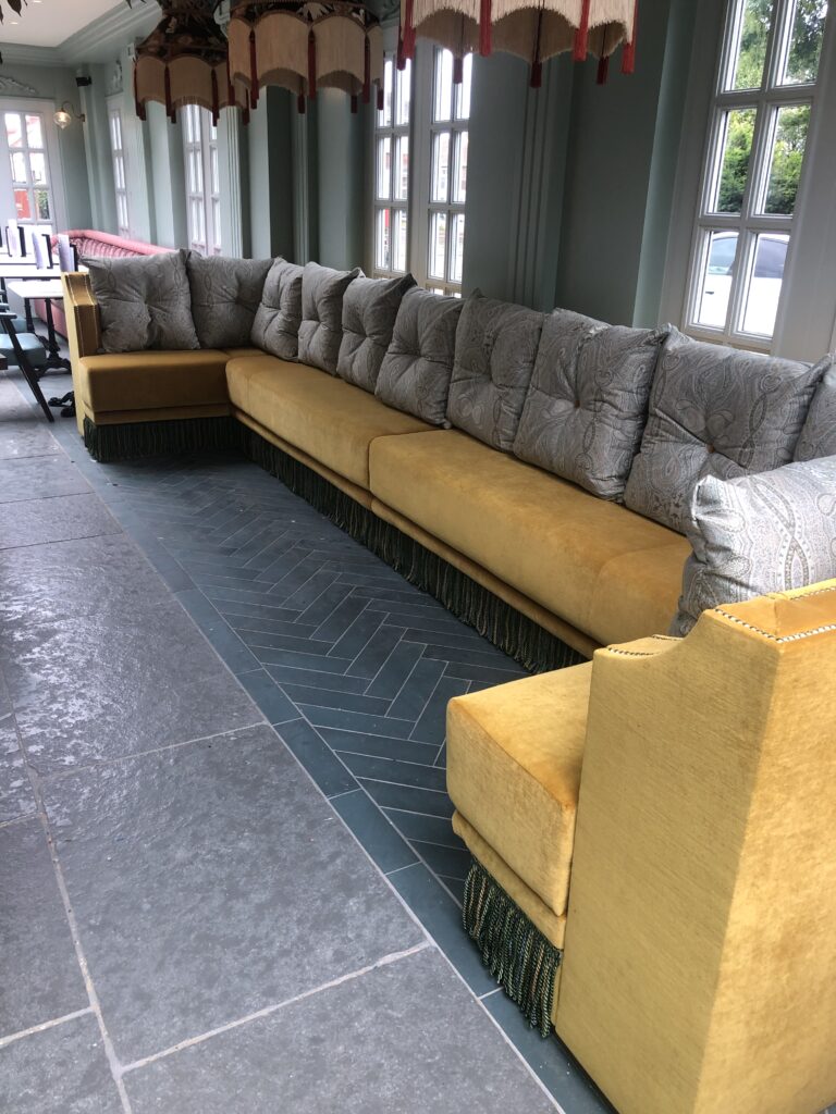 Mustard corner banquette with grey patterned back cushions, stud edging and green fringe.