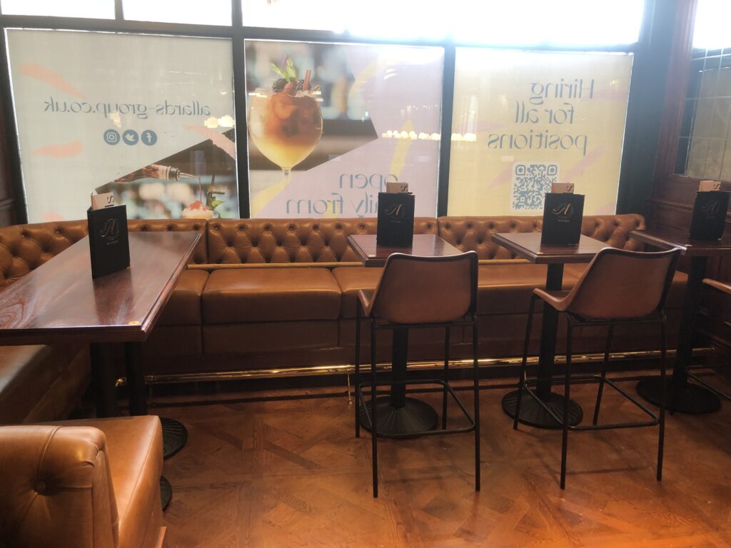 Tan buttoned banquette by a window with timber tables and a brass-coloured footrail.