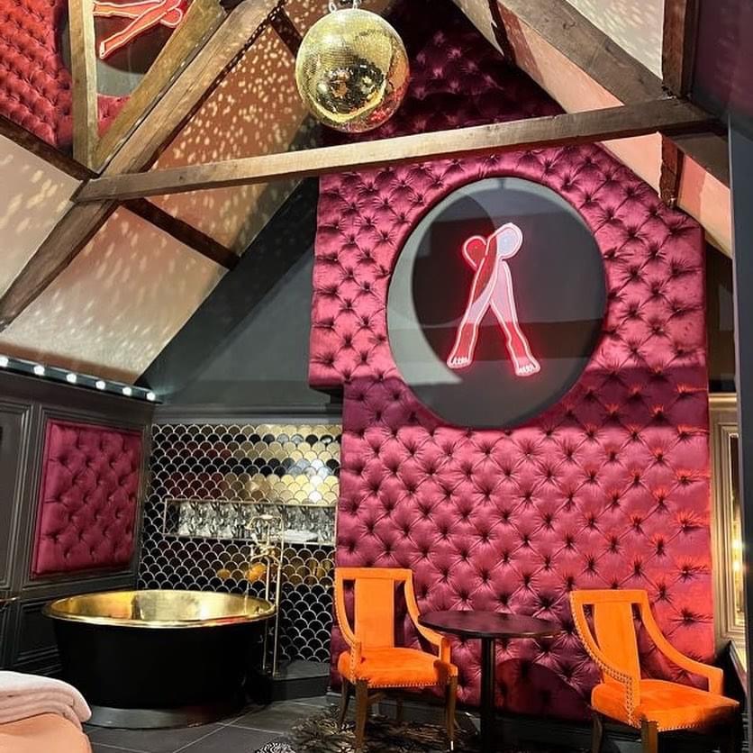 Hotel interior with tall burgundy buttoned wall upholstery, a circular neon feature, timber beams and a metallic bath.