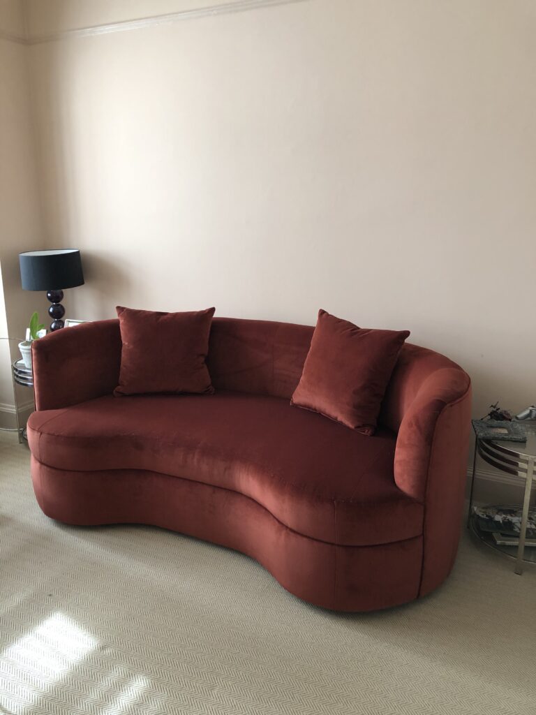 Rust-coloured sofa with a curved front, rounded arms and two matching scatter cushions in a light room.
