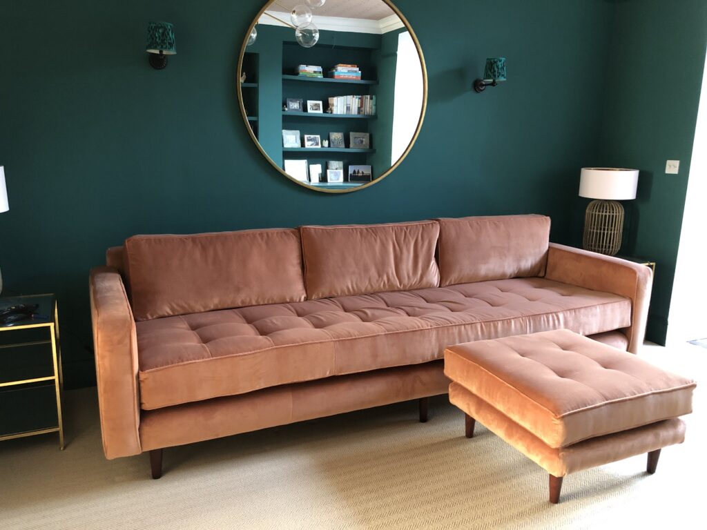 Rose-coloured sofa with a buttoned seat, three back cushions and a matching footstool against a dark green wall.