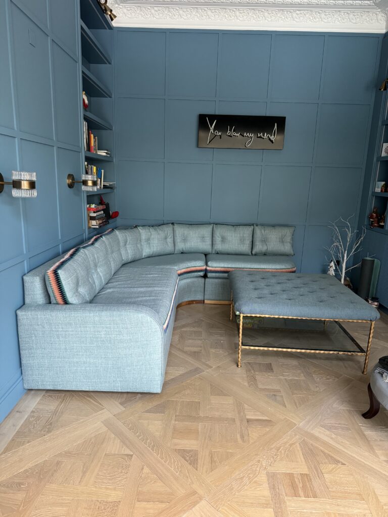 Blue-grey corner seating and a buttoned footstool in a room with blue panelling and decorative white cornicing.