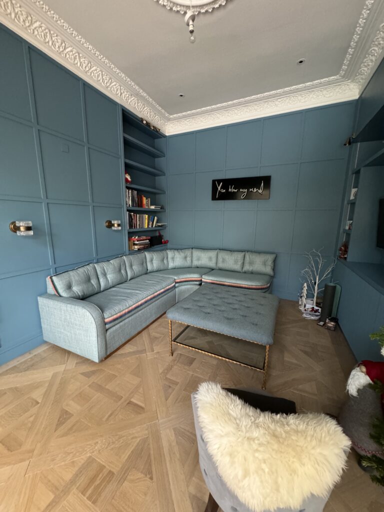 Wide portrait view of the blue-panelled room, corner sofa, buttoned footstool and herringbone floor.