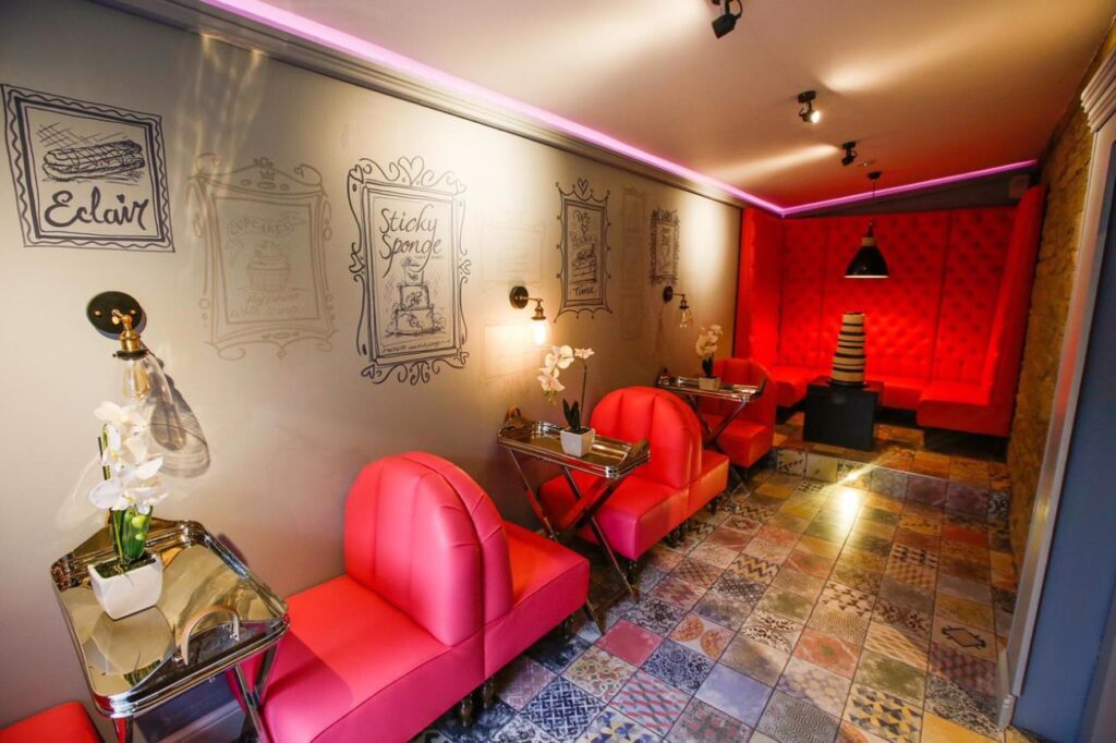 Wide view of rounded pink chairs, metal tray tables and a pink banquette at the far end of a tiled room.