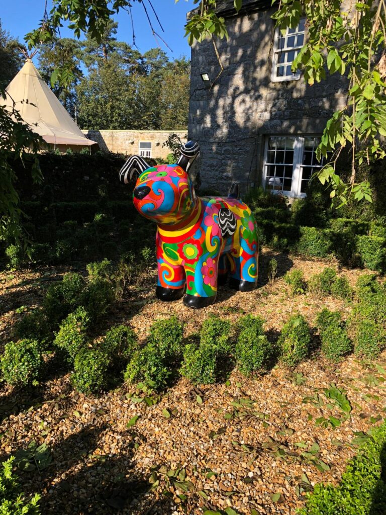 Colourfully painted dog sculpture among clipped hedges beside Woodhill Hall’s stone building.