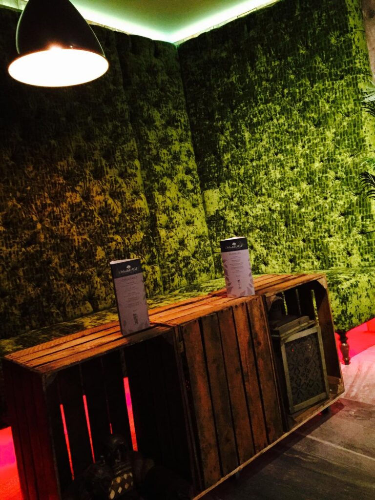 High-backed green buttoned banquette behind wooden crate tables and a black pendant light.