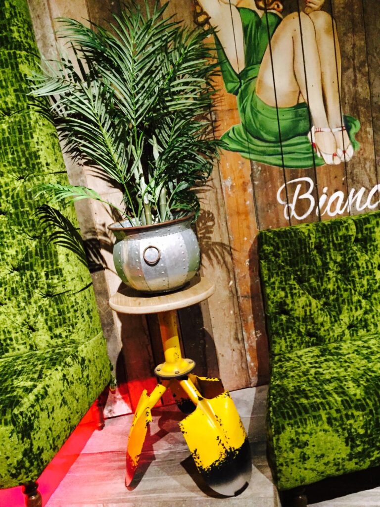 Green upholstered seats beside a metal planter on a yellow shovel-legged stand.