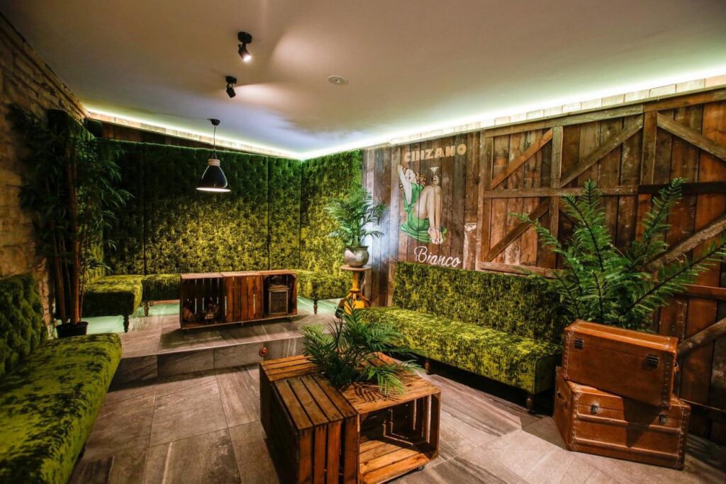 Wide view of green banquette seating, a matching sofa, timber walls, crate tables and plants.
